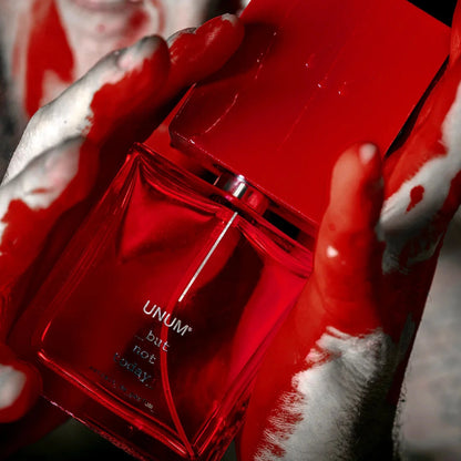 Person with red and white makeup holding a red perfume bottle labeled 'UNUM'.